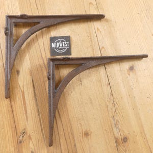 May include: Two L-shaped shelf brackets made of dark brown cast iron with a textured finish. Each bracket features a triangular support brace and pre-drilled holes for mounting. A small square black tag with the white text MIDWEST is included.