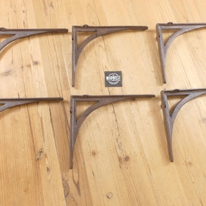 May include: A set of six brown cast iron shelf brackets arranged in two rows of three. Each bracket features a triangular support structure with a curved brace. A small square black label with the white text MIDWEST is centered between them.