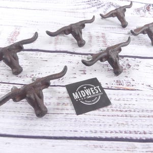 May include: Six cast iron bull head cabinet pulls with a rustic brown finish. The pulls are arranged on a white wooden surface. A black tag with the text "MIDWEST CRAFT HOUSE" is also present.