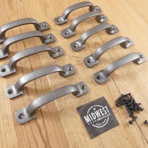 May include: A set of eight grey metal drawer pulls with a textured finish, each featuring two screw holes at the ends. A small square black label with the white text MIDWEST CRAFT FORGE and a small pile of black screws are included in the set.