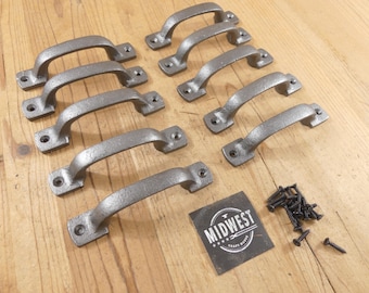 May include: A set of eight grey metal drawer pulls with a textured finish, each featuring two screw holes at the ends. A small square black label with the white text MIDWEST CRAFT FORGE and a small pile of black screws are included in the set.