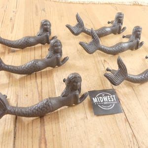May include: A set of five dark gray cast iron drawer pulls shaped like mermaids, each featuring a textured tail and a reclining figure. A small black square tag with the white text MIDWEST CRAFT HOUSE is positioned among the hardware pieces.