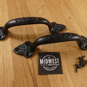 May include: Two black, ornate cast iron handles with decorative ends and a curved grip. The handles are accompanied by a small black label with the text "MIDWEST CRAFT HOUSE" and a small pile of screws. The handles are on a wooden surface.