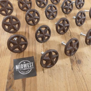 May include: A collection of rustic brown cast iron star-shaped cabinet knobs. The knobs have a five-pointed star design within a circular frame. A black square label with the text "MIDWEST" is also visible.