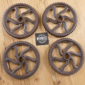 May include: Four antique cast iron gears with a rusty brown finish. Each gear has a central hub with six curved spokes and a toothed outer rim. A black and white label with the text "MIDWEST" is between the gears.