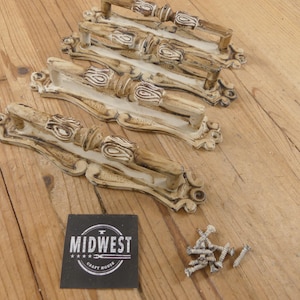 May include: Four ornate, cream-colored door handles with a distressed finish, featuring detailed carvings and a handle. A small pile of screws and a black label with the text "MIDWEST CRAFT HOUSE" are also present.