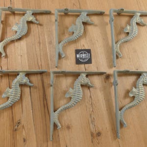 May include: Six cast iron seahorse shelf brackets in a distressed, light gray finish. Each bracket features a seahorse design and a right-angle shape for mounting. The brackets are arranged on a wooden surface.