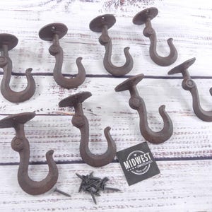 May include: A collection of eight rustic brown cast iron ceiling hooks with mounting plates. The hooks are curved and have a decorative design. A small pile of screws and a tag with the text "MIDWEST CRAFT SUPPLY CO." are also present.