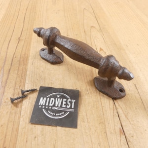 May include: A dark brown cast iron door handle with a textured, rustic finish and two mounting points. Beside the handle are two small black screws and a square black card featuring the white text MIDWEST CRAFT HOUSE with a small anvil icon.