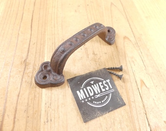 May include: A dark brown, textured cast iron handle with a curved grip featuring raised circular studs and decorative, clover-shaped mounting plates at each end. Two black metal screws are included, alongside a square black card with the white text MIDWEST CRAFT HOUSE and a small anvil icon.