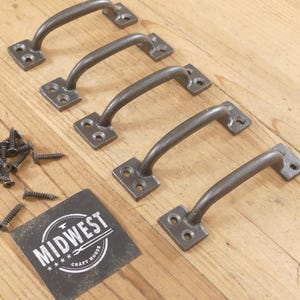 May include: Five dark gray metal cabinet pulls with matching screws are arranged on a light wood surface. Each pull has a rectangular base with two screw holes and a curved handle. A black and white label reads "MIDWEST CRAFT HOUSE."