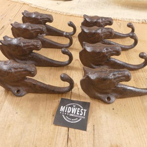 May include: Six dark brown cast iron wall hooks, each featuring a sculpted horse head design with a curved hook at the base. A small square black sign with white text reading MIDWEST CRAFT HOUSE is positioned in the center of the arrangement.
