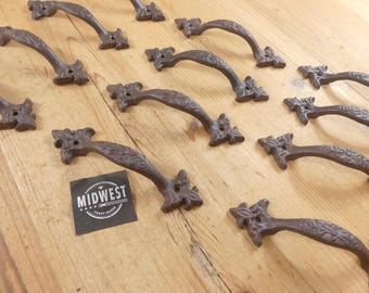 May include: A collection of dark brown cast iron drawer pulls featuring an embossed floral pattern and two screw holes at each end. A small black square label with the white text MIDWEST CRAFT HOUSE is positioned next to one of the handles.