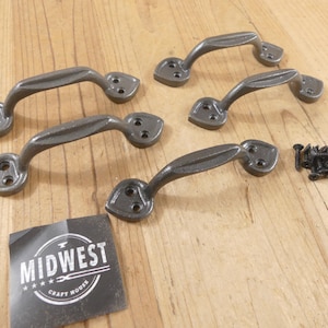 May include: Five dark gray metal handles with a curved design, each with two mounting points. A small pile of black screws is next to the handles. A black and white label with the text "MIDWEST CRAFT HOUSE" is also present.