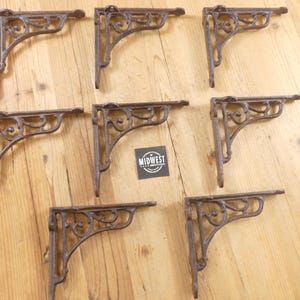 May include: Eight brown cast iron shelf brackets featuring an ornate scrollwork design with mounting holes at the ends. A small square black sign with the white text MIDWEST is positioned in the center among the arranged metal brackets.