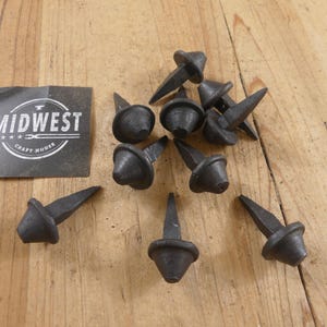 May include: A collection of nine dark gray, hand-forged iron nails with conical heads and square-tapered shanks. A square black label with white text reading MIDWEST CRAFT HOUSE is positioned to the left of the metal hardware pieces.