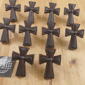 May include: Ten dark brown cast iron cross-shaped drawer knobs with a textured finish. Each knob features a central point and flared arms. A small square black tag with the white text MIDWEST CRAFT HOUSE is positioned in the lower left corner.