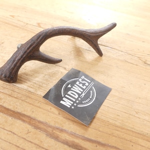 May include: A dark brown, textured metal cabinet pull shaped like a deer antler. Next to the pull is a square black sticker with white text that reads MIDWEST CRAFT HOUSE, featuring a small graphic of a hammer and anvil between the words.