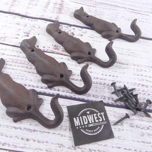 May include: Four cast iron dog-shaped hooks with curved tails, in a rustic brown finish. Includes a pile of black screws and a black label with white text that reads "MIDWEST CRAFT HOUSE".