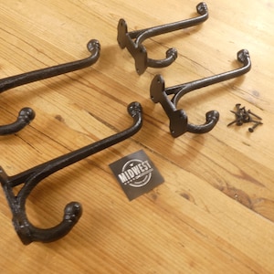 May include: Four black cast iron coat hooks with a curved design, displayed on a wooden surface. The hooks are accompanied by mounting screws and a small tag with the word "MIDWEST" on it.