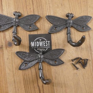 May include: Three cast iron dragonfly hooks with a dark gray finish. Each hook features detailed wing designs and a curved hook. The word "MIDWEST" is printed on a black card. Mounting screws are included.