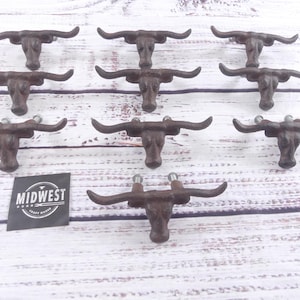 May include: Nine cast iron bull head cabinet pulls with a rustic brown finish. The pulls are arranged on a white wooden surface. A black and white Midwest Craft House logo is in the lower left corner.