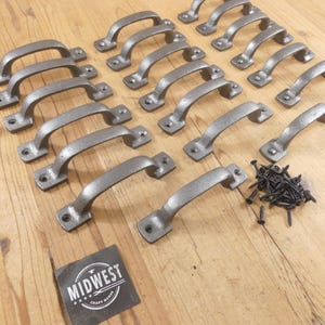 May include: A collection of fifteen grey metal drawer pulls with a textured finish, each featuring two screw holes at the ends. A small pile of black screws is included, along with a square black label featuring the text MIDWEST CRAFT HOUSE.