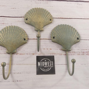 May include: Three seashell-shaped wall hooks with a distressed, aged finish. The hooks are a muted green-gold color and have a curved hook at the bottom. A black square with white text reading "MIDWEST CRAFT HOUSE" is also present.