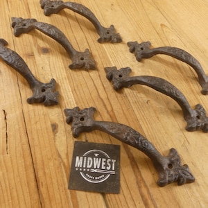 May include: Set of six cast iron door handles with an ornate, floral design. The handles are dark brown and have a rustic, antique appearance. The handles are arranged on a wooden surface, with a small tag that reads "Midwest Craft House".