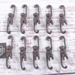 May include: A collection of eleven cast iron monkey-shaped hooks. The hooks are a dark brown color and are arranged on a light-colored wooden surface. Each hook features a monkey hanging from a curved hook.