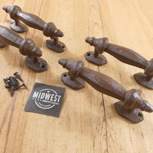 May include: Four dark brown cast iron door handles with a textured, rustic finish. Each handle features a central grip with decorative ends and two mounting plates with screw holes. A small black square card displays the white text MIDWEST CRAFT MOVES.