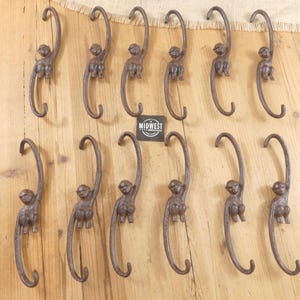May include: Twelve brown cast iron S-hooks, each featuring a sculpted monkey figure integrated into the center of the hook. A small black square tag with the white text MIDWEST is positioned in the center of the arrangement of these metal hooks.