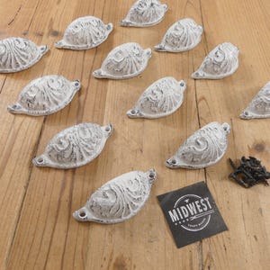 May include: A collection of ten white, distressed-finish metal cup pulls with ornate, raised scrollwork patterns. Each pull features two screw holes for mounting. A small black square card with the white text MIDWEST CRAFT HOUSE is included.