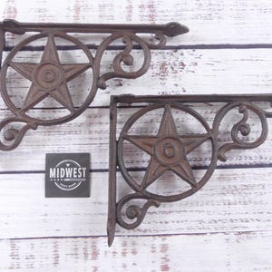 May include: Two rustic, brown cast iron shelf brackets. Each bracket features a star within a circle, with decorative scrollwork. The brackets are designed to be mounted on a wall and support a shelf. The image also includes a small sign that says "Midwest".