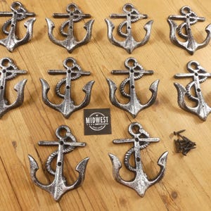 May include: A collection of ten silver-colored cast iron anchor wall hooks with rope detailing. The hooks are arranged on a wooden surface, with a small black square that reads "MIDWEST".