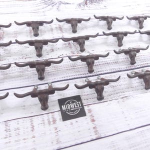 May include: A collection of cast iron bull head cabinet pulls with long horns. The pulls are a dark brown color and are arranged on a white wooden surface. A black and white Midwest Craft Supply logo is visible.
