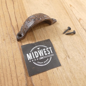 May include: A dark brown, textured cast iron cup pull handle with two matching black screws. A square black label is present with the white text MIDWEST CRAFT HOUSE and a graphic of an anvil and tongs inside a circular border design.