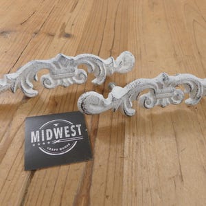 May include: Two distressed white decorative drawer pulls with ornate scrollwork and a dark gray accent. The pulls are on a wooden surface with a black card that reads "MIDWEST CRAFT HOUSE".