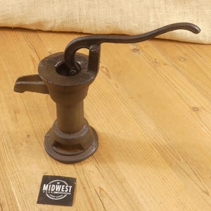 May include: A vintage cast iron water pump with a curved handle. The pump is dark brown and sits on a wooden surface. The pump has a spout and a round base. A small black and white Midwest Crafty House sticker is in the foreground.