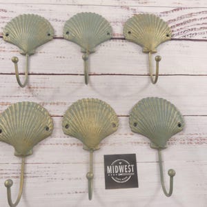 May include: Six seashell-shaped wall hooks in a seafoam green color with a metallic gold finish. Each hook has a curved, downward-facing hook. The hooks are arranged on a light-colored, wood-grain surface. A black square with the text "MIDWEST" is also present.