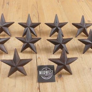 May include: Ten rustic, dark brown cast iron star-shaped decorative objects. The stars are arranged on a light brown wooden surface. A black and white label with the text "MIDWEST CRAFT HOUSE" is also present.