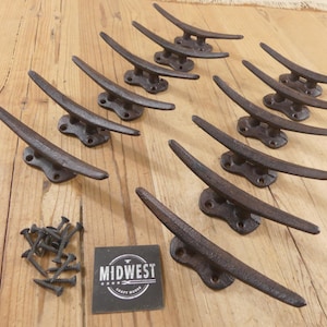 May include: A collection of ten cast iron cleats, perfect for nautical decor. The cleats are dark brown with a weathered finish, arranged on a light wood surface. A pile of screws and a black label with the text "MIDWEST CRAFT HOUSE" are also present.