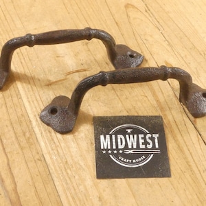 May include: Two rustic, dark brown cast iron handles with a textured finish. Each handle has a curved bar and two mounting plates with holes. A small square sign with the text "MIDWEST CRAFT HOUSE" is also present.