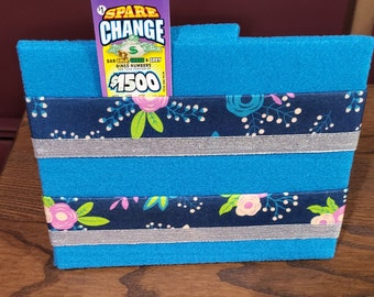 Può includere: Una scatola di feltro blu con due strati di tessuto floreale blu scuro e bianco. La scatola ha un biglietto della lotteria in cima che dice "Spare Change" e "1500 €".