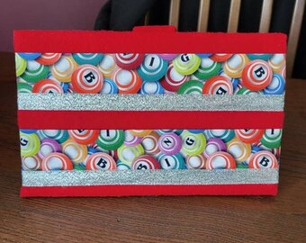 Può includere: Porta carte da bingo in feltro rosso con due strisce di glitter argentate. Il porta carte presenta un motivo colorato di palline da bingo con lettere e numeri.