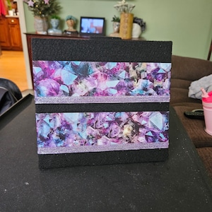 May include: A black square-shaped box with two strips of purple and blue patterned tape with silver glitter accents. The tape features a design of purple, blue, and white crystals.