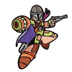 May include: An embroidered patch featuring a cartoon-style Mandalorian character in a green and brown outfit, holding a blaster and jetpack. The character is wearing a helmet with a visor and a purple cape.