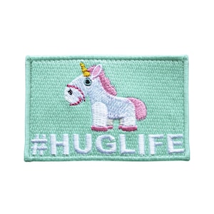 Puede incluir: Un parche bordado de color verde claro con un unicornio blanco con una crin rosa y un cuerno amarillo. El unicornio está de pie sobre un fondo verde claro con el texto "#HUGLIFE" en blanco.