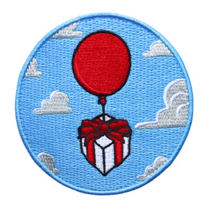 Puede incluir: Un globo rojo con una caja de regalo blanca y roja atada a él, flotando en un cielo azul con nubes blancas. El globo y la caja de regalo están bordados en un parche circular azul.
