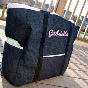 May include: A denim diaper bag with pink trim and a white pocket. The bag has black straps and the name "Gabriella" embroidered in pink on the front.